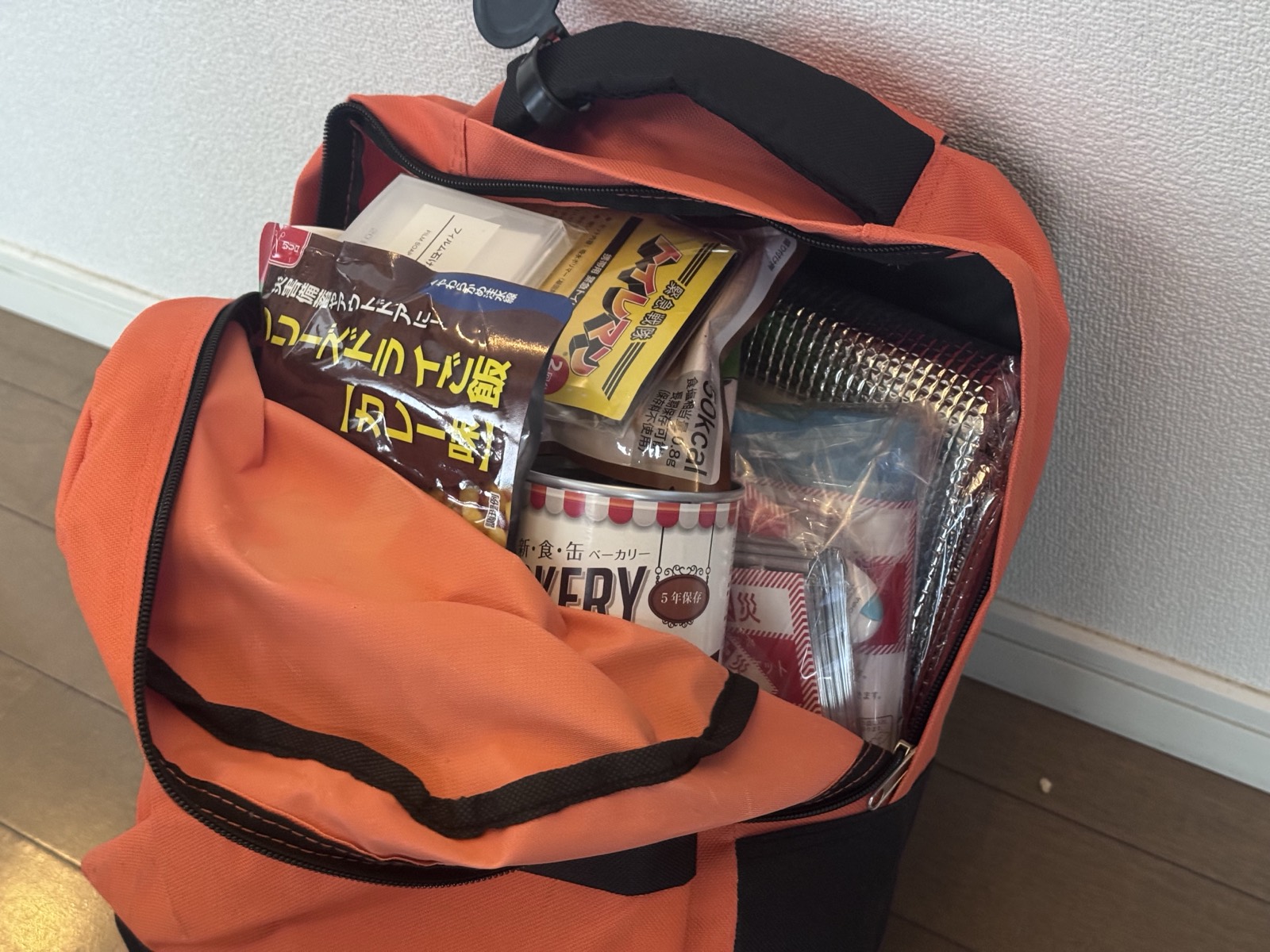 オレンジ色の防災リュックを開けた状態。アルファ米のカレー、5年保存の缶パン、簡易トイレ、アルミの保温シート、救急用品などが入っている